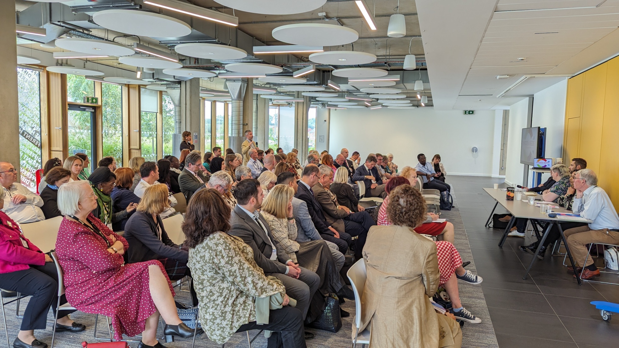 A seated audience attending the Northamptonshire Hidden Needs Report launch event, with one audience member standing to ask a question to the seated panel members at the front of the room